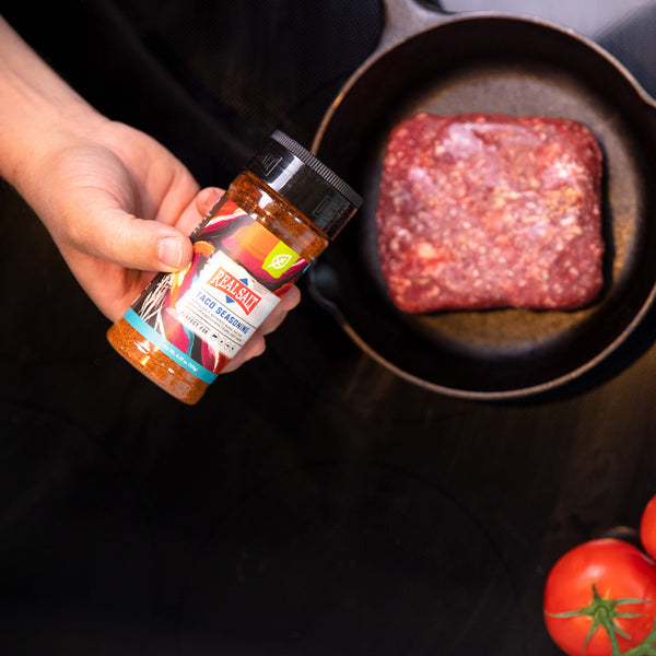 Hand holds a bottle of Real Salt Taco Seasoning over a cast iron skillet with ground beef browning.