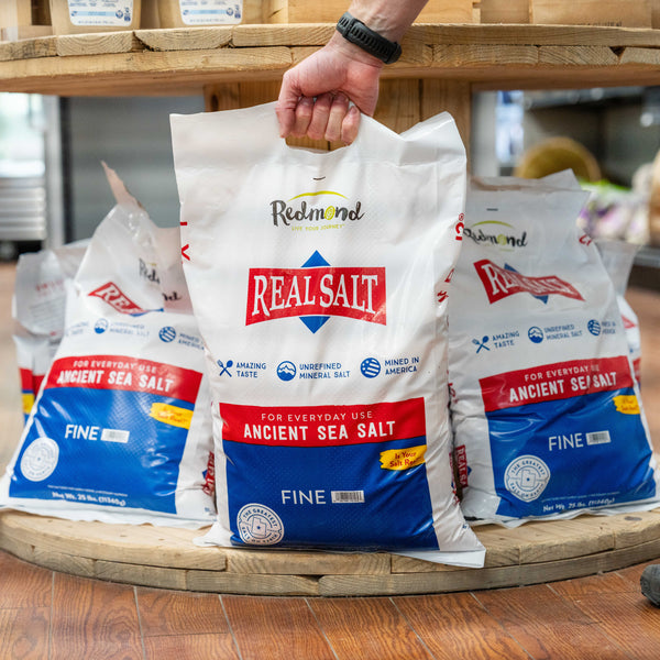 Hand lifts a 25lb bag of Redmond Real Salt Ancient Sea Salt from a wooden display.