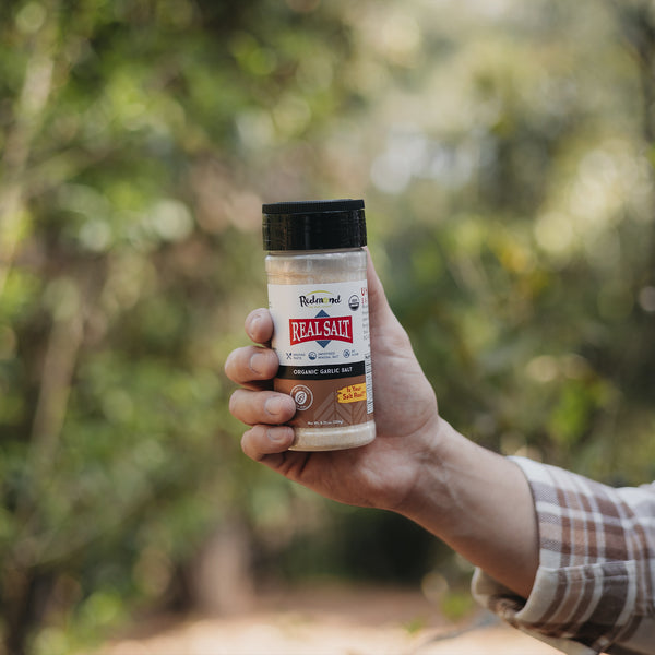 Hand holds a shaker of Redmond Real Salt organic garlic salt outdoors with blurred greenery in the background.