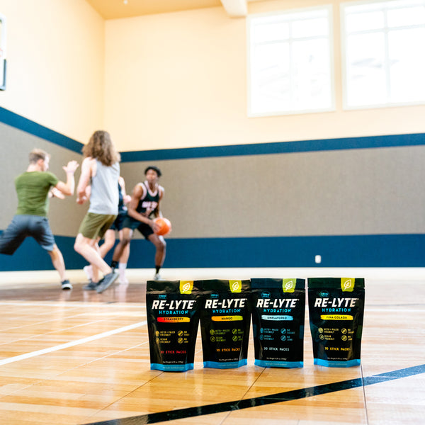 Four Re-Lyte Hydration 30 count stick packs electrolyte powder on a basketball court with young men playing basketball in the background. Flavors from left to right: Strawberry Lemonade, Mango, Unflavored and Piña Colada.