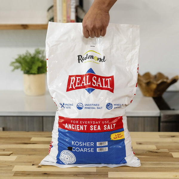 Hand holds a large bag of Redmond Real Salt, an unrefined, kosher, ancient sea salt mined in America. 
