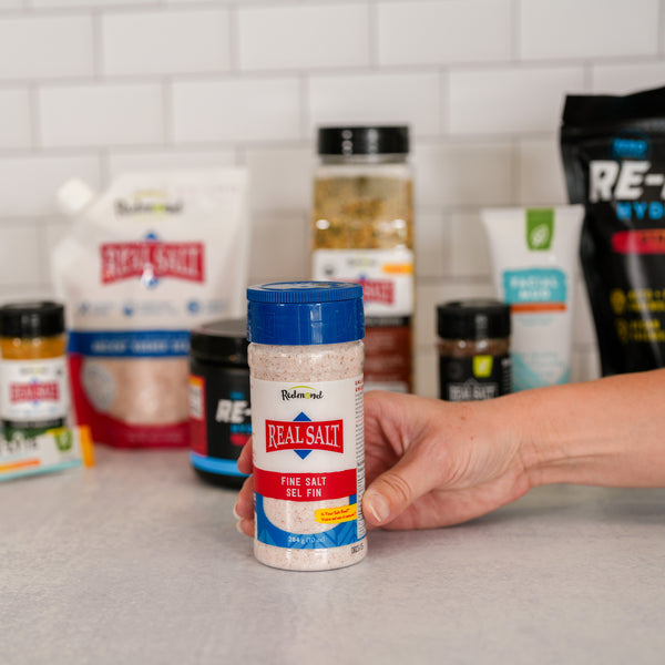 Hand holds a shaker of Redmond Real Salt fine salt against a backdrop of other Redmond Real Salt products.