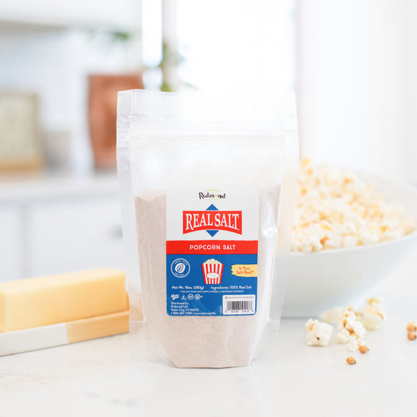 A bag of Redmond Real Salt Popcorn Salt stands on a countertop, ready to season a bowl of fresh popcorn. 
